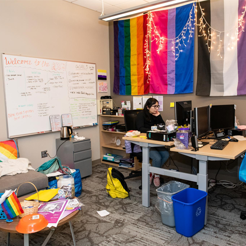 student looking at a computer in the quuer resource center with multiple pride flags and lights hanging in the room