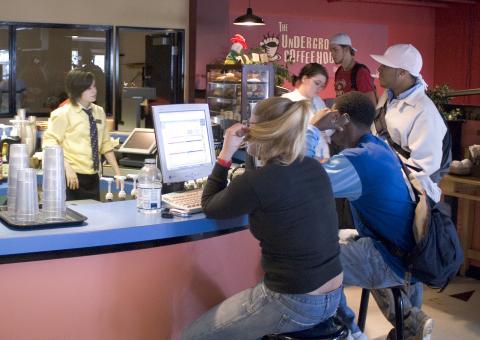 An image of students standing at the coffee counter, one is working on a computer stationed there